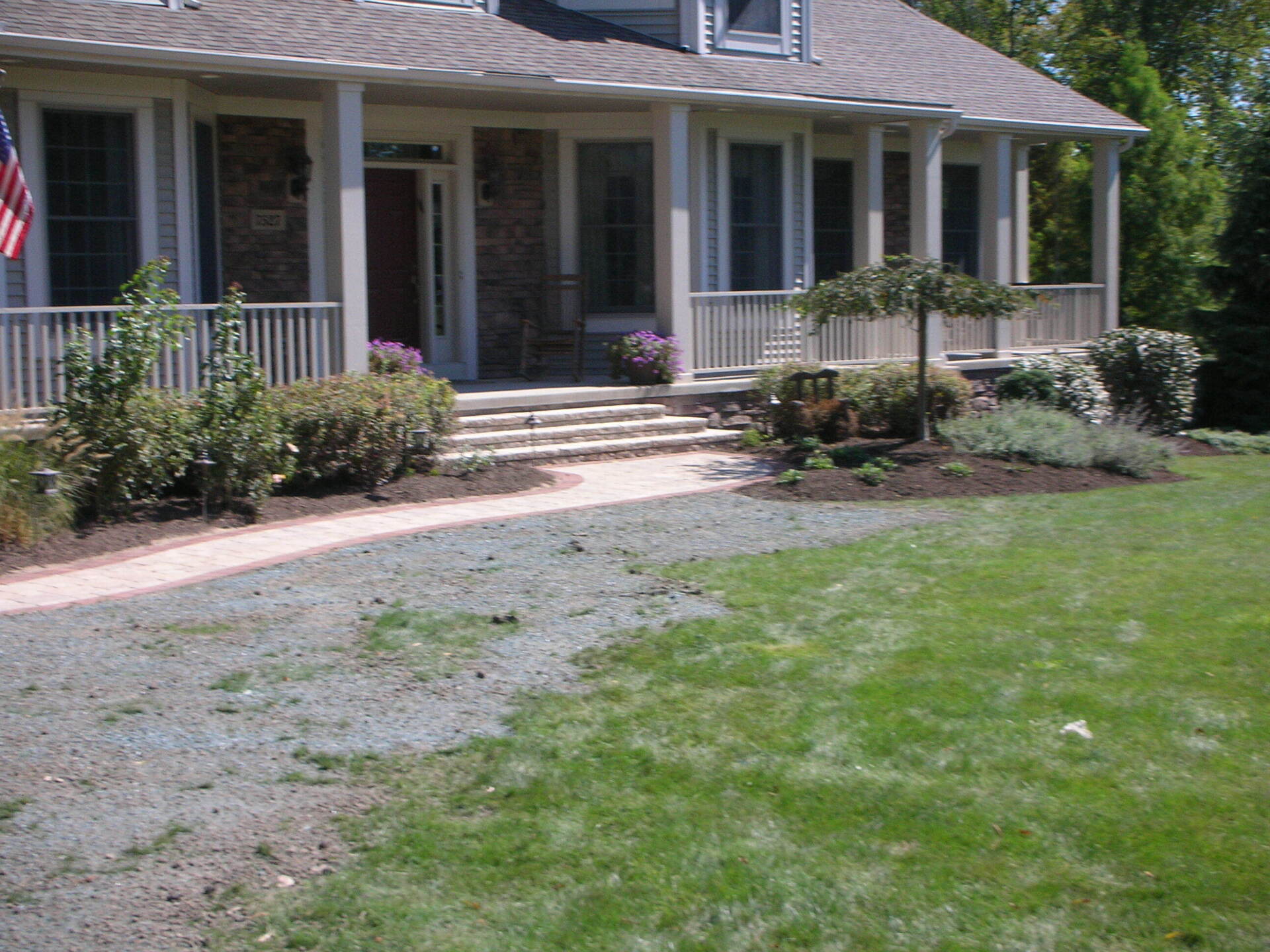 Front porch of a house with white columns, shrubs, and a gravel driveway beside a green lawn.