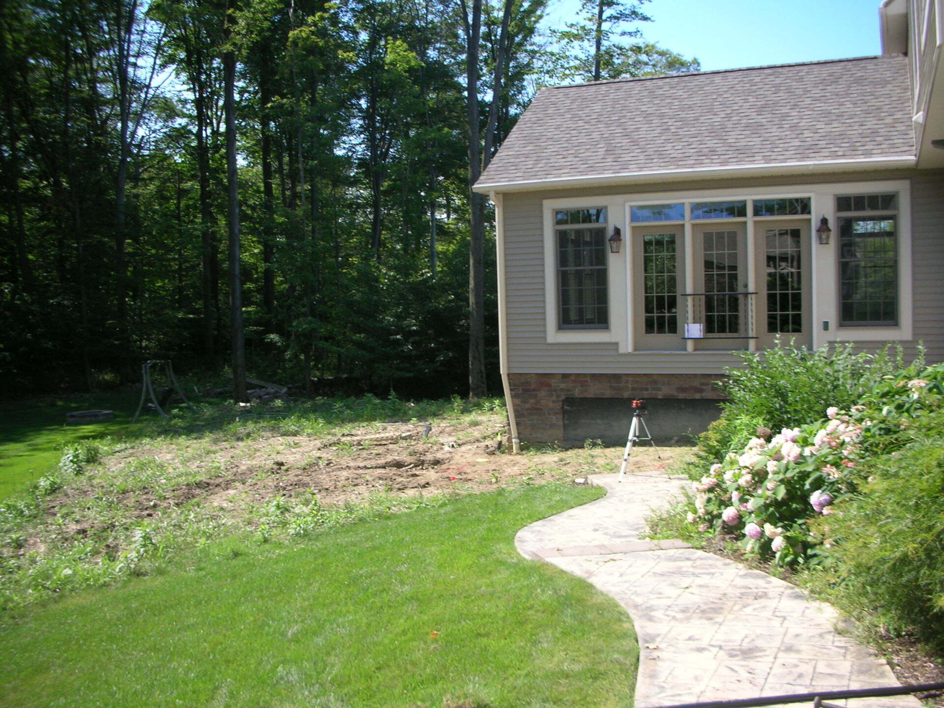 Small house exterior with a curved stone walkway, lawn, trees, and flowering shrubs.