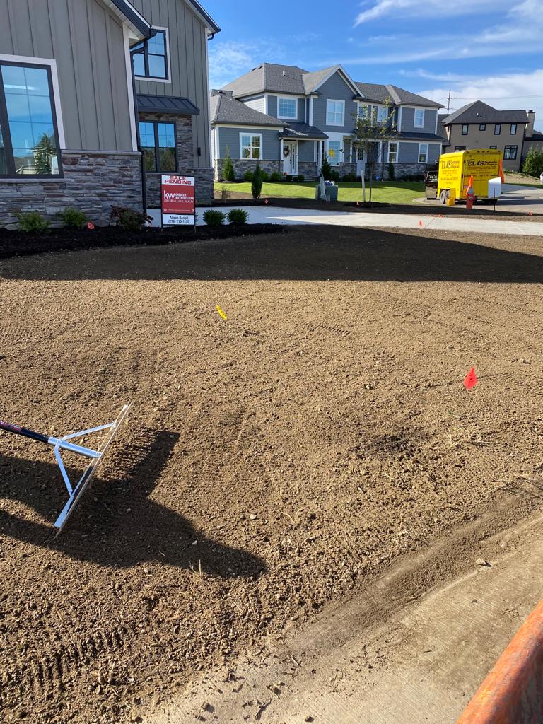Fresh gravel being spread in a suburban front yard beside new houses and a parked yellow machine.