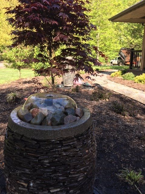 Backyard stone fountain beside a burgundy tree and patio in bright sunlight