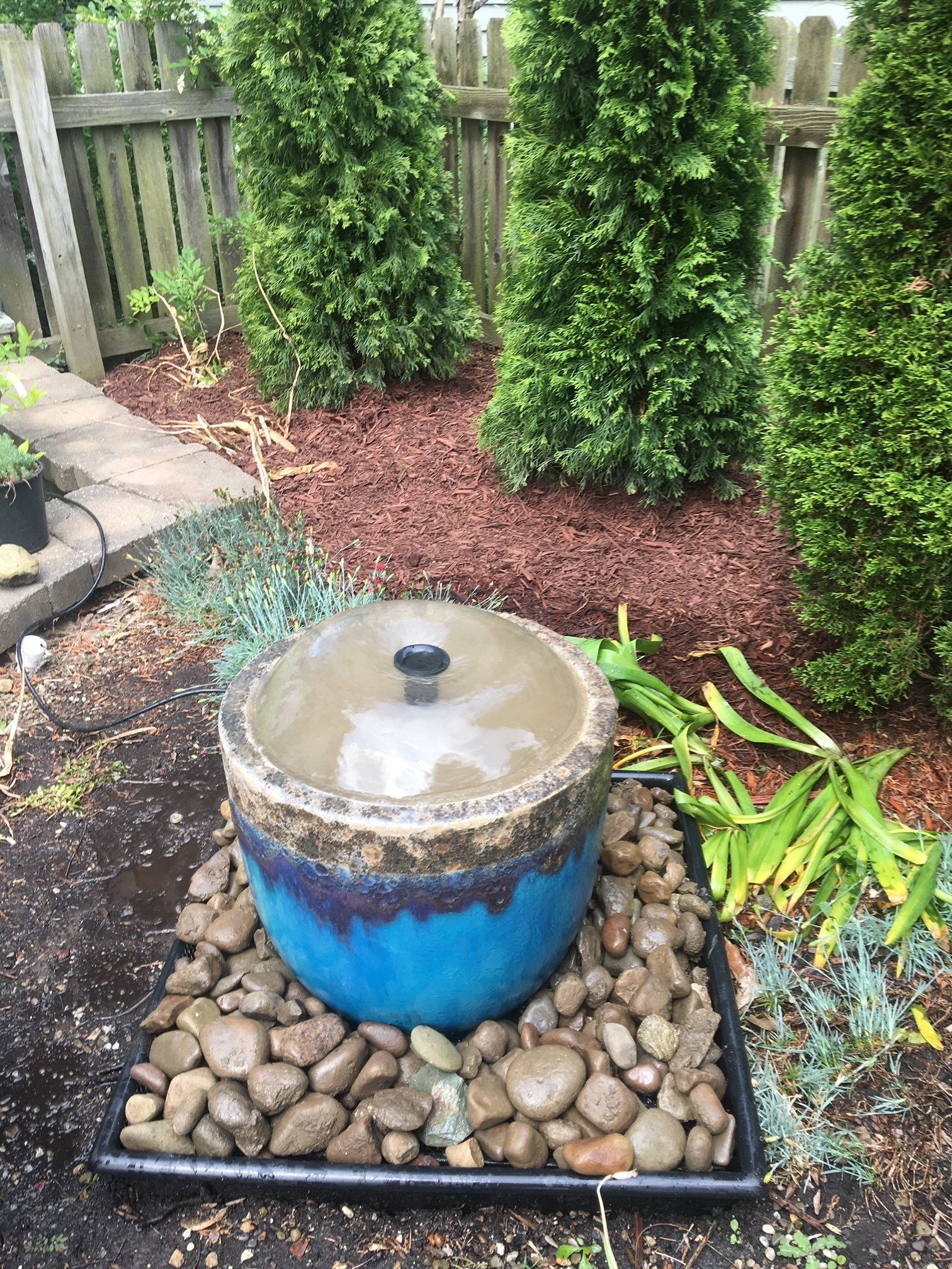 Blue backyard fountain bubbling in a stone basin beside rocks, mulch, and green shrubs