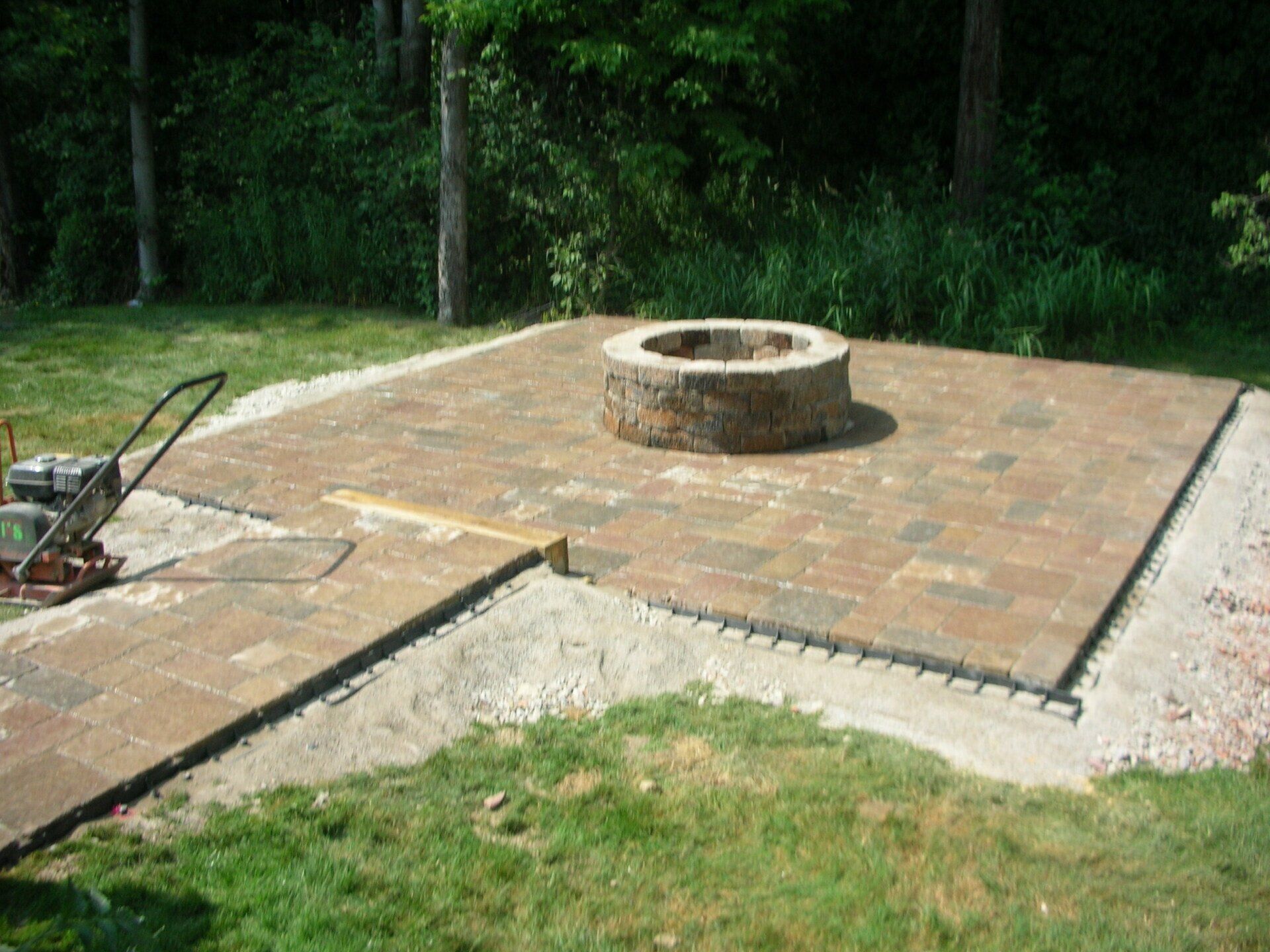 Brick patio with a circular stone fire pit in a grassy backyard, bordered by trees.