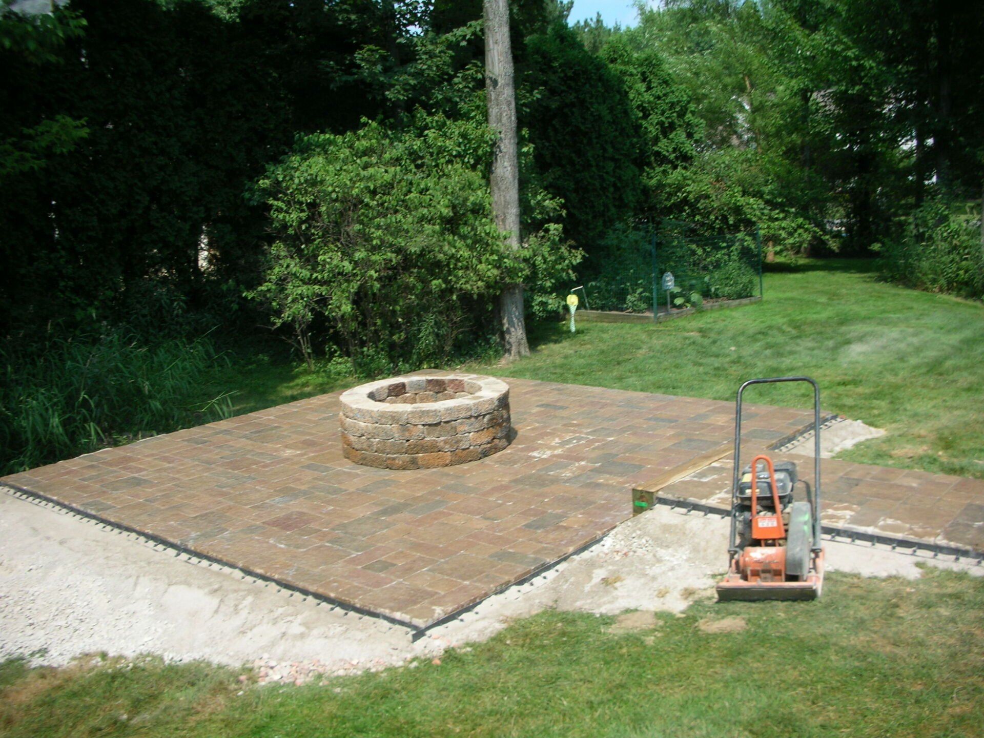 Brick patio with a circular fire pit beside a grassy yard and an orange saw at the edge