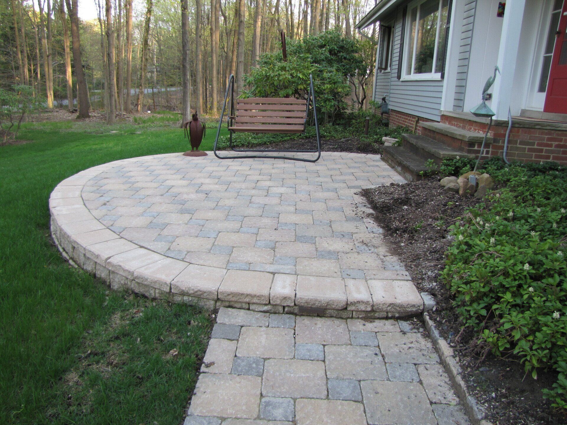 Curved stone patio with bench and fire pit beside a house, bordered by grass and trees