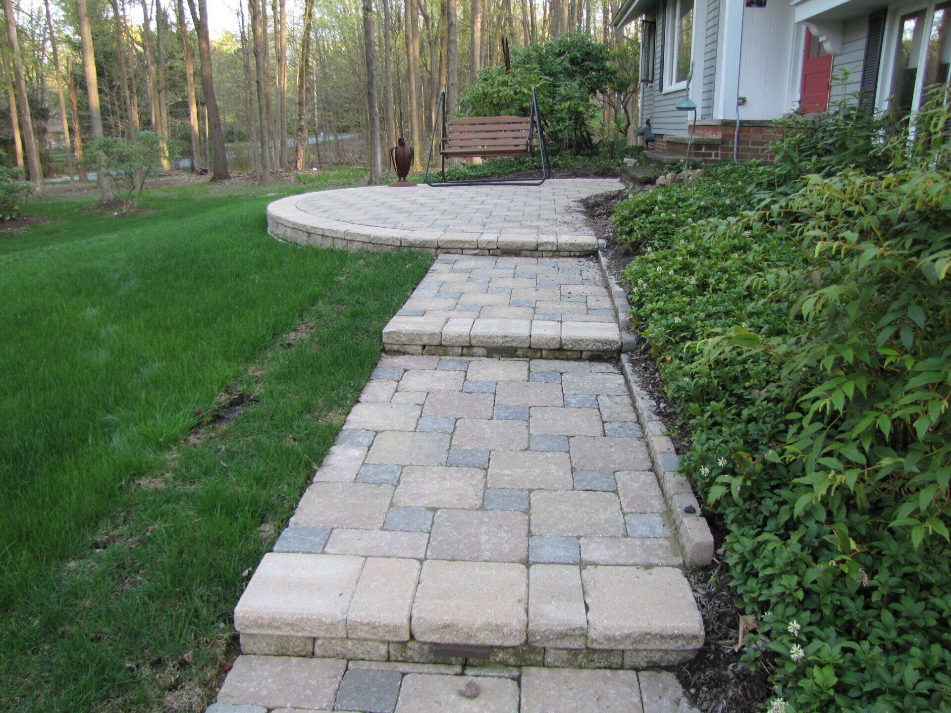 Stone walkway through green yard leading to a house and bench in a wooded garden
