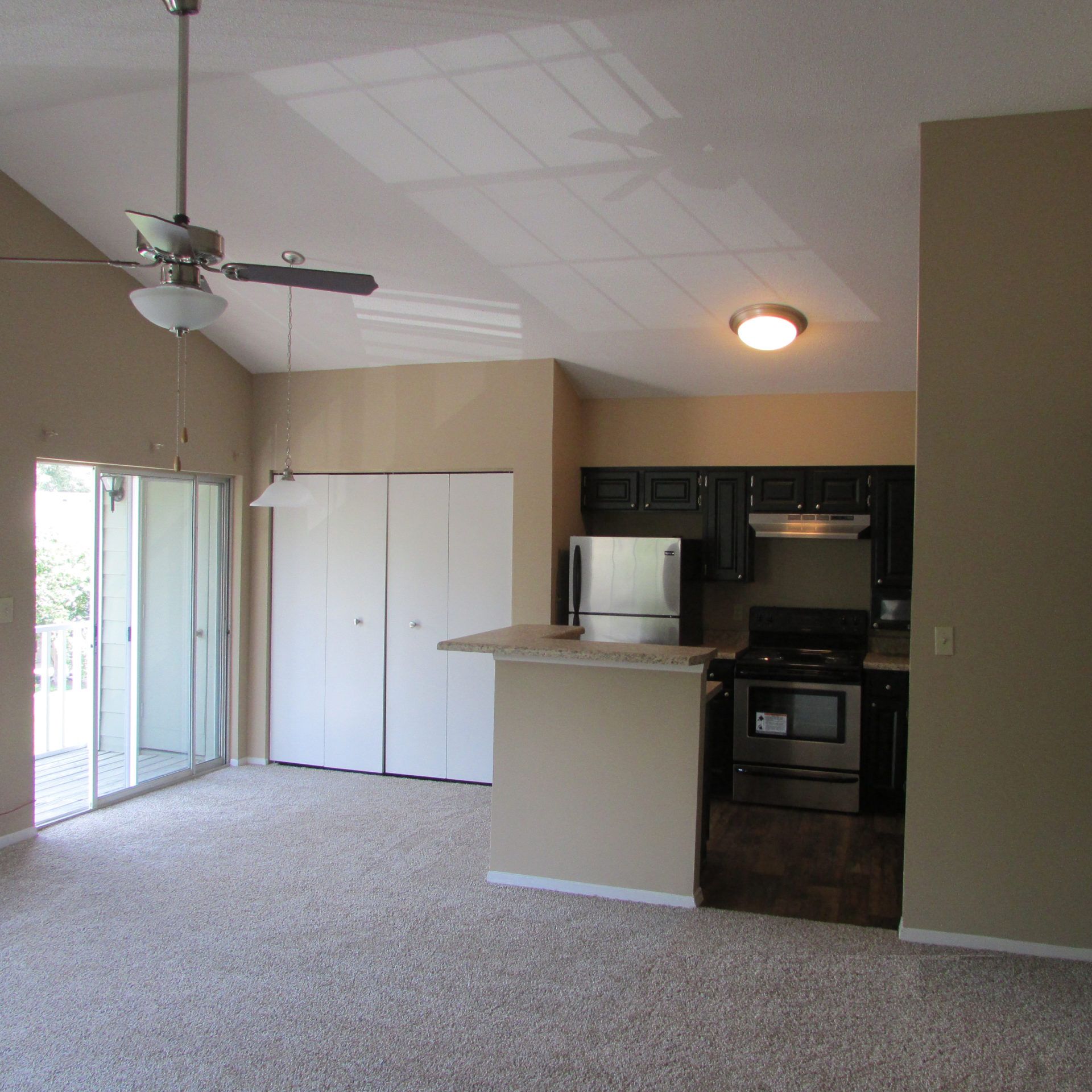 Empty living room with beige carpet, vaulted ceiling, ceiling fan, and small kitchen area.