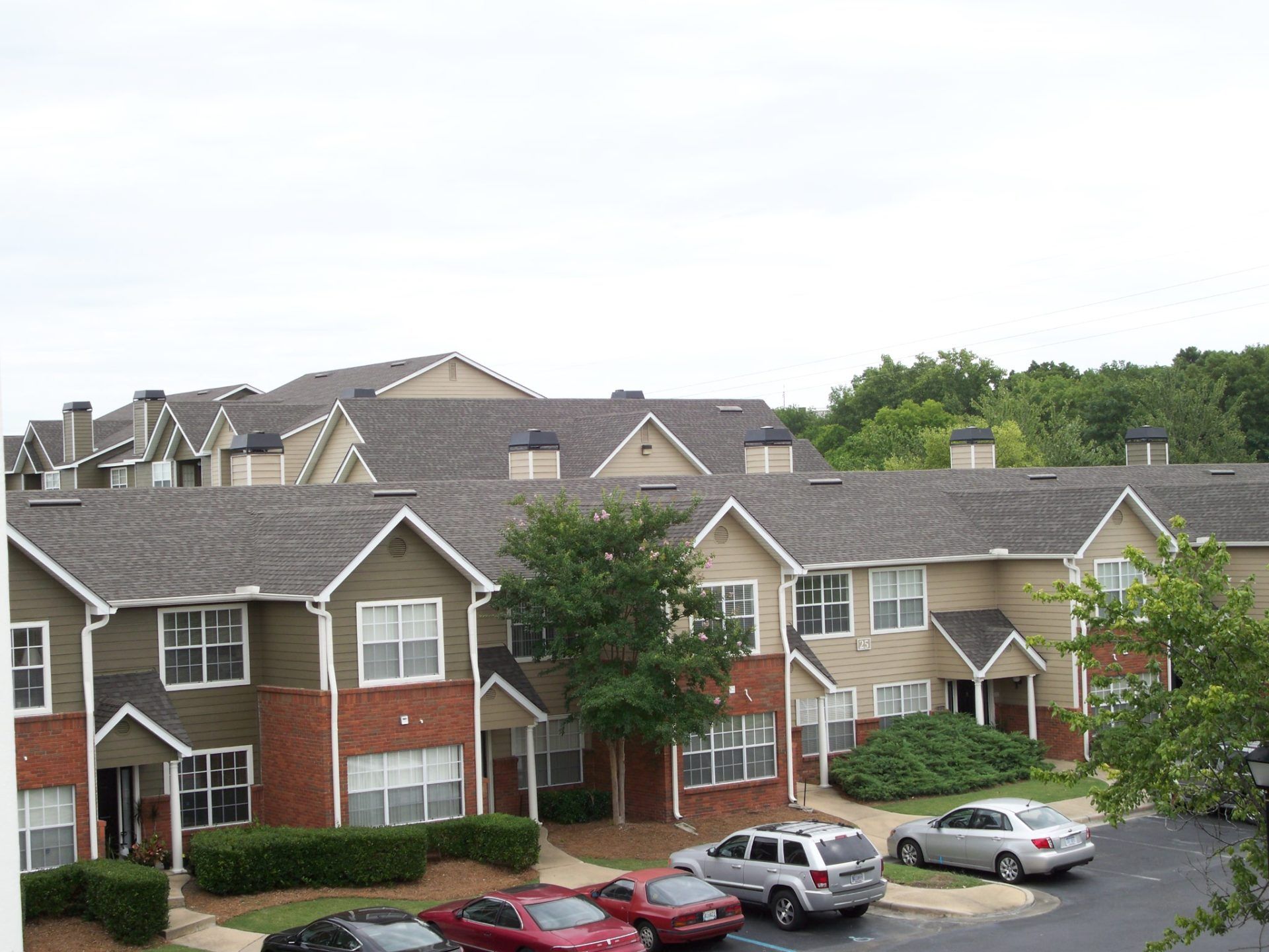 Apartment complex with brick and beige townhomes, parked cars, and trees under a cloudy sky
