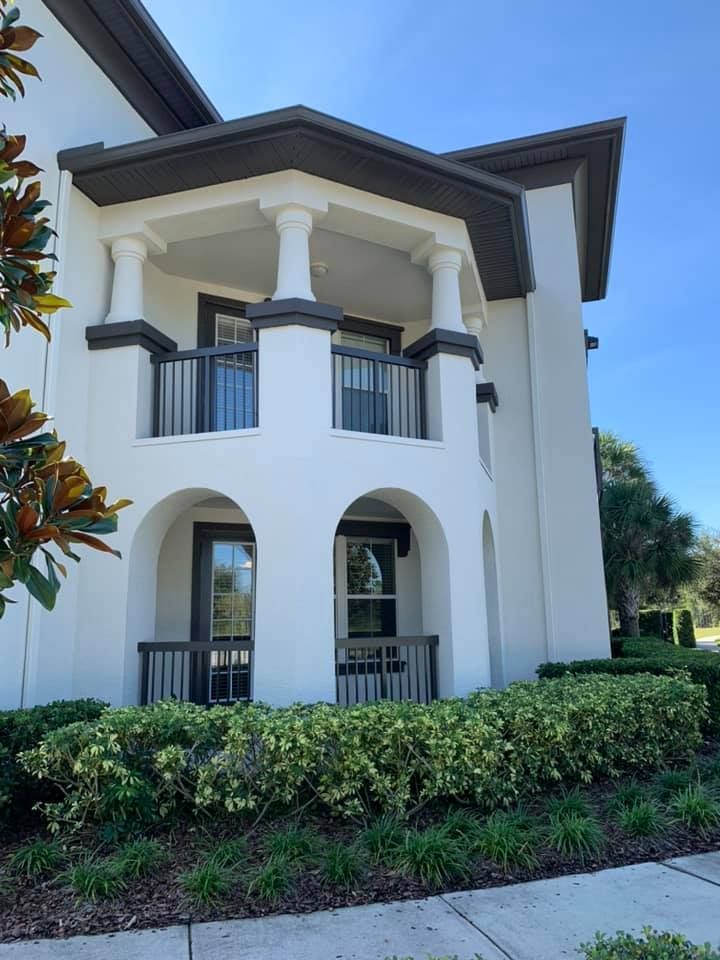 White two-story Mediterranean-style house with arched balconies, dark roof, and green shrubs in front