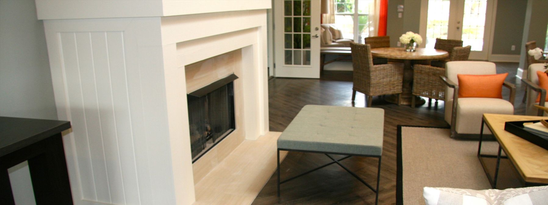 Bright living room with a white fireplace, beige rug, and orange-accented seating near tall windows.
