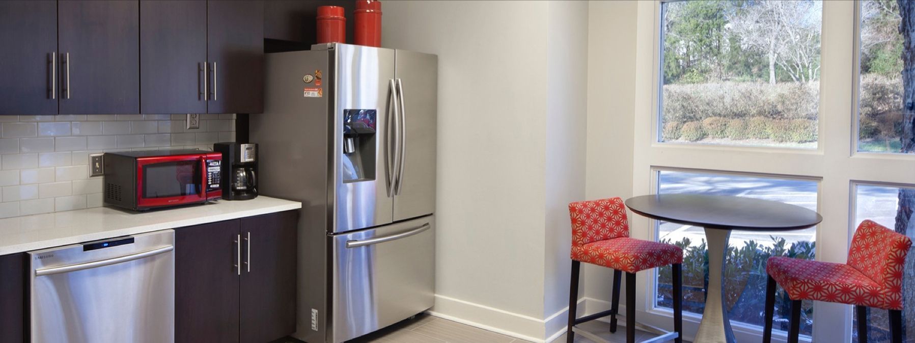 Modern kitchen with dark cabinets, stainless fridge, red microwave, and a small table by bright windows