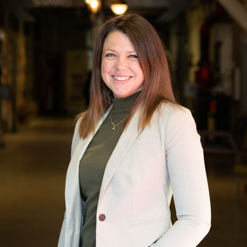 Woman with brown hair smiles, wearing a green turtleneck and beige blazer in an indoor hallway.