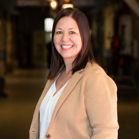 Woman with shoulder-length dark hair, smiling, wearing a tan blazer and white blouse, in a hallway.