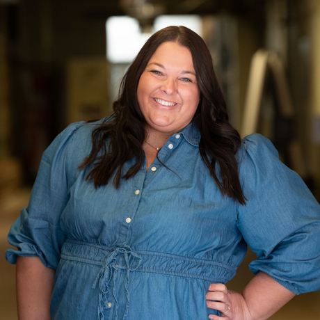 Woman with dark hair, smiling, wearing a blue denim dress, hand on hip.