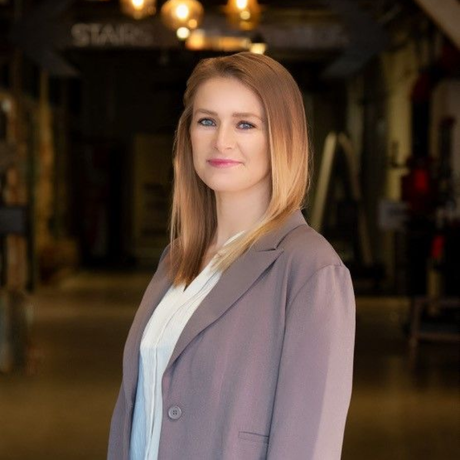 Woman in a mauve blazer, white shirt, and blonde hair, smiling in an industrial-chic hallway.