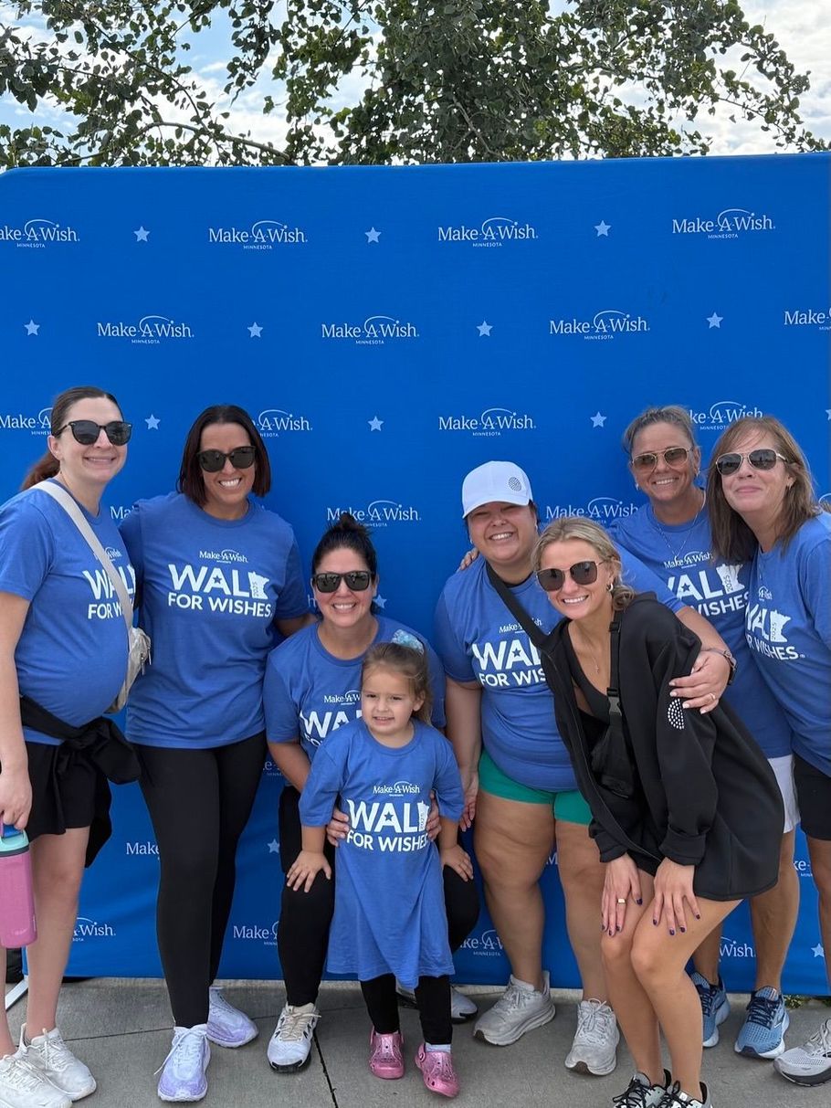 Group of eight people smiling in blue "Walk for Wishes" t-shirts, in front of a blue backdrop outdoors.