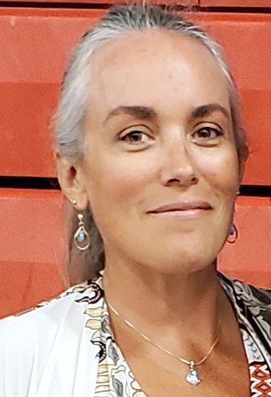 Woman with gray hair, smiling, wearing earrings and a necklace, in front of an orange background.