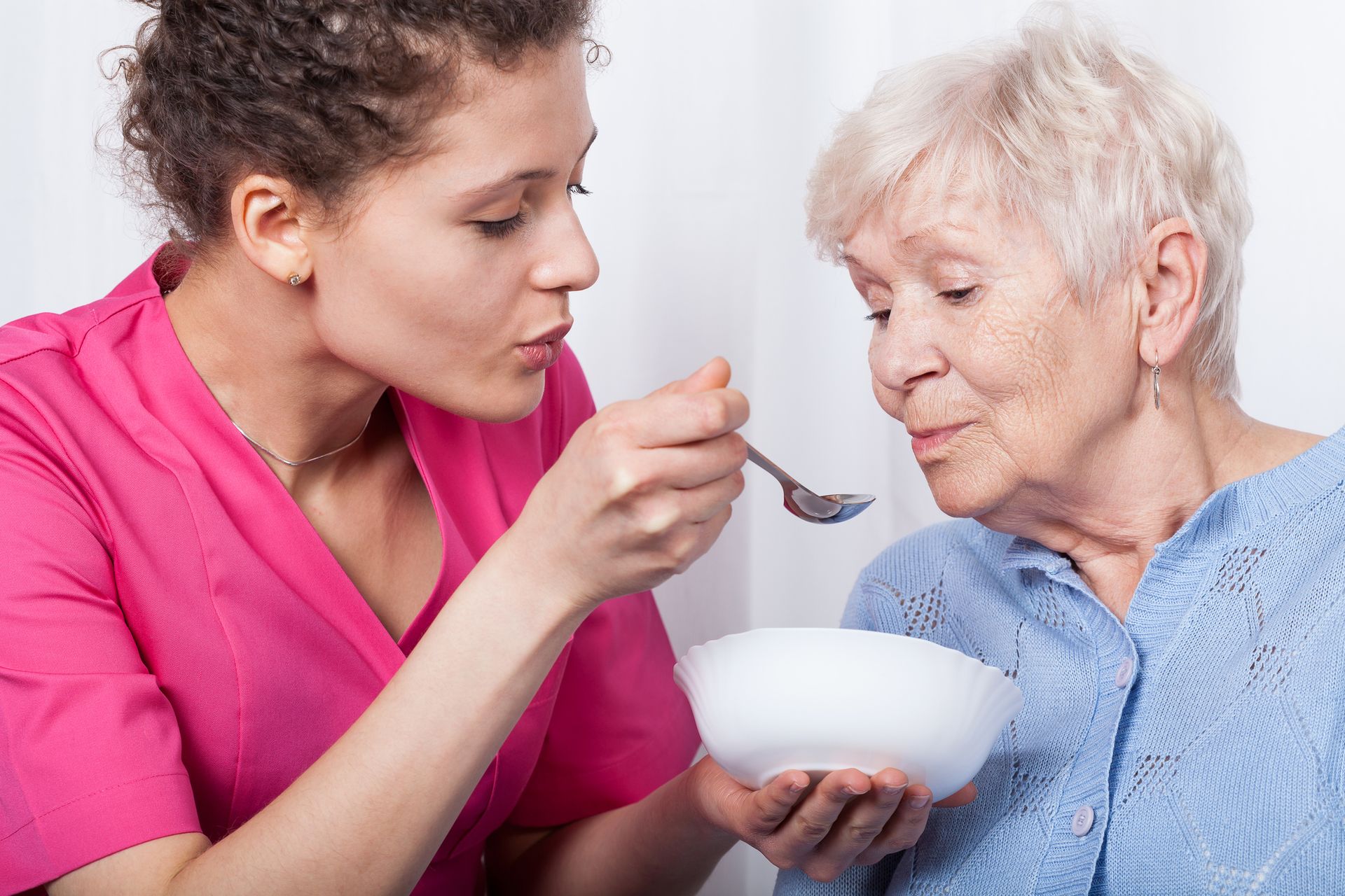 Female nurse in pink scrubs feeds senior woman with spoon, showcasing palliative care services.