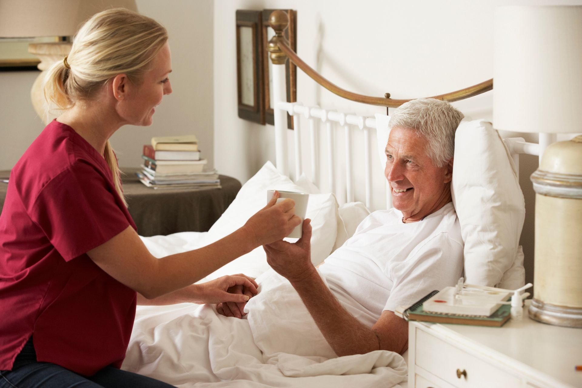 Woman in red scrubs gives senior male a cup as he lays in bed, showcasing palliative care services.