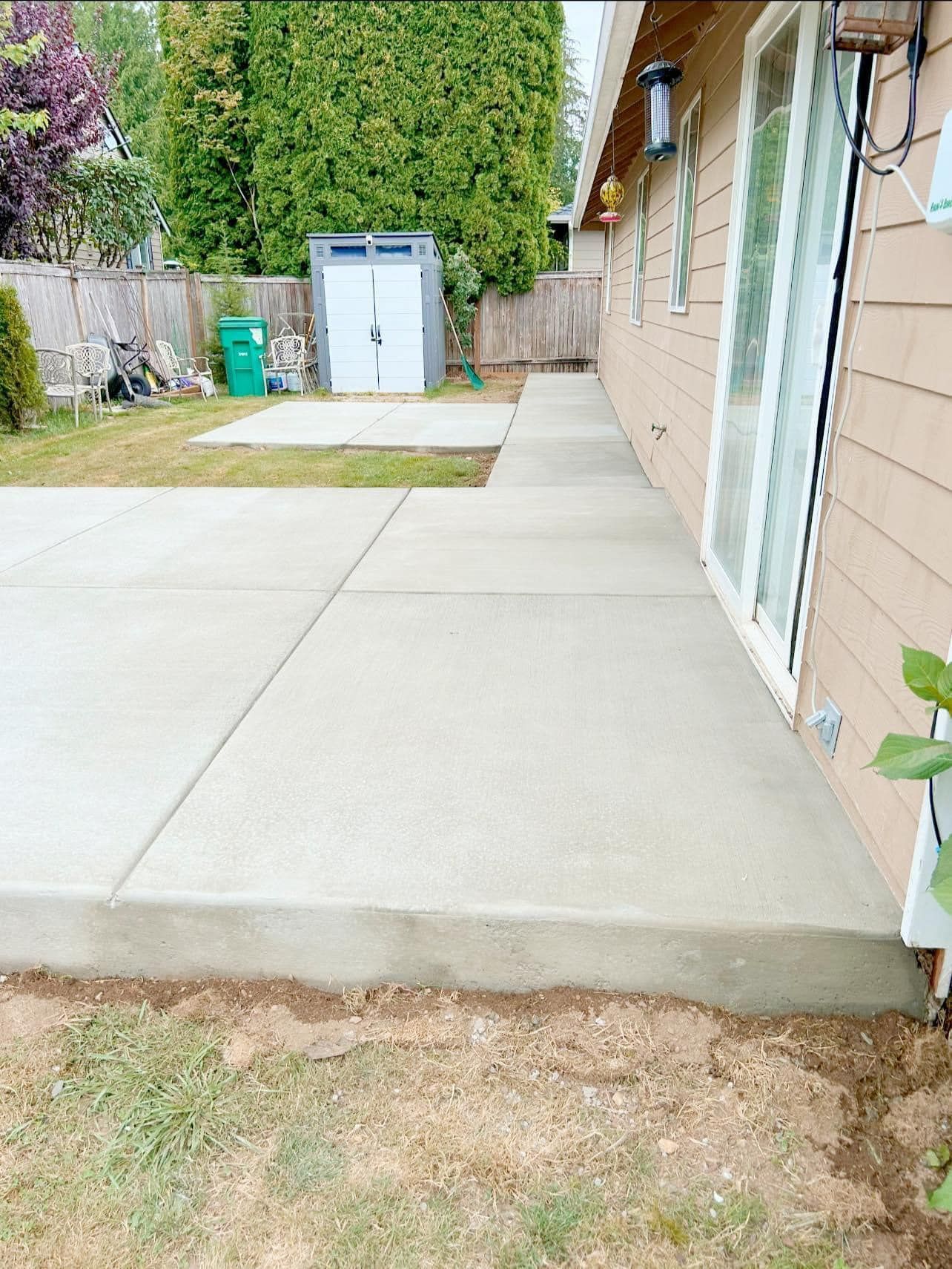 Concrete patio next to a house with a sliding glass door. Grass and a shed are in the background.
