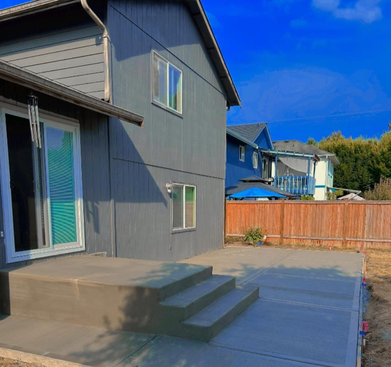 Backyard of a two-story gray house with concrete patio and steps, wooden fence, and blue sky.