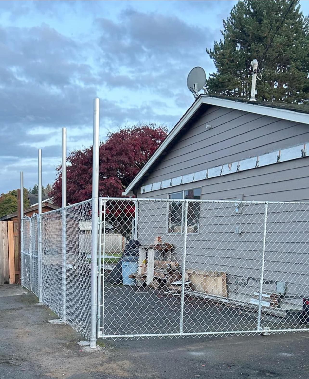 A chain-link fence surrounds a house and yard, with tall posts. Overcast sky.