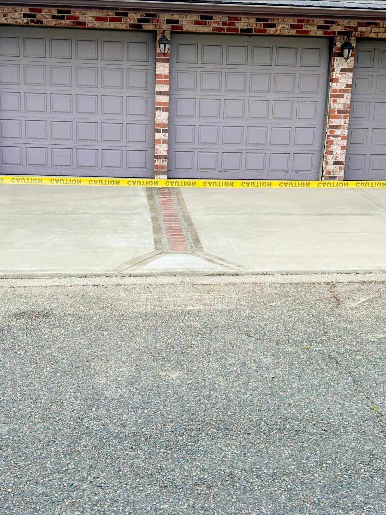 Three gray garage doors with yellow caution tape, brick accents, and a concrete driveway.