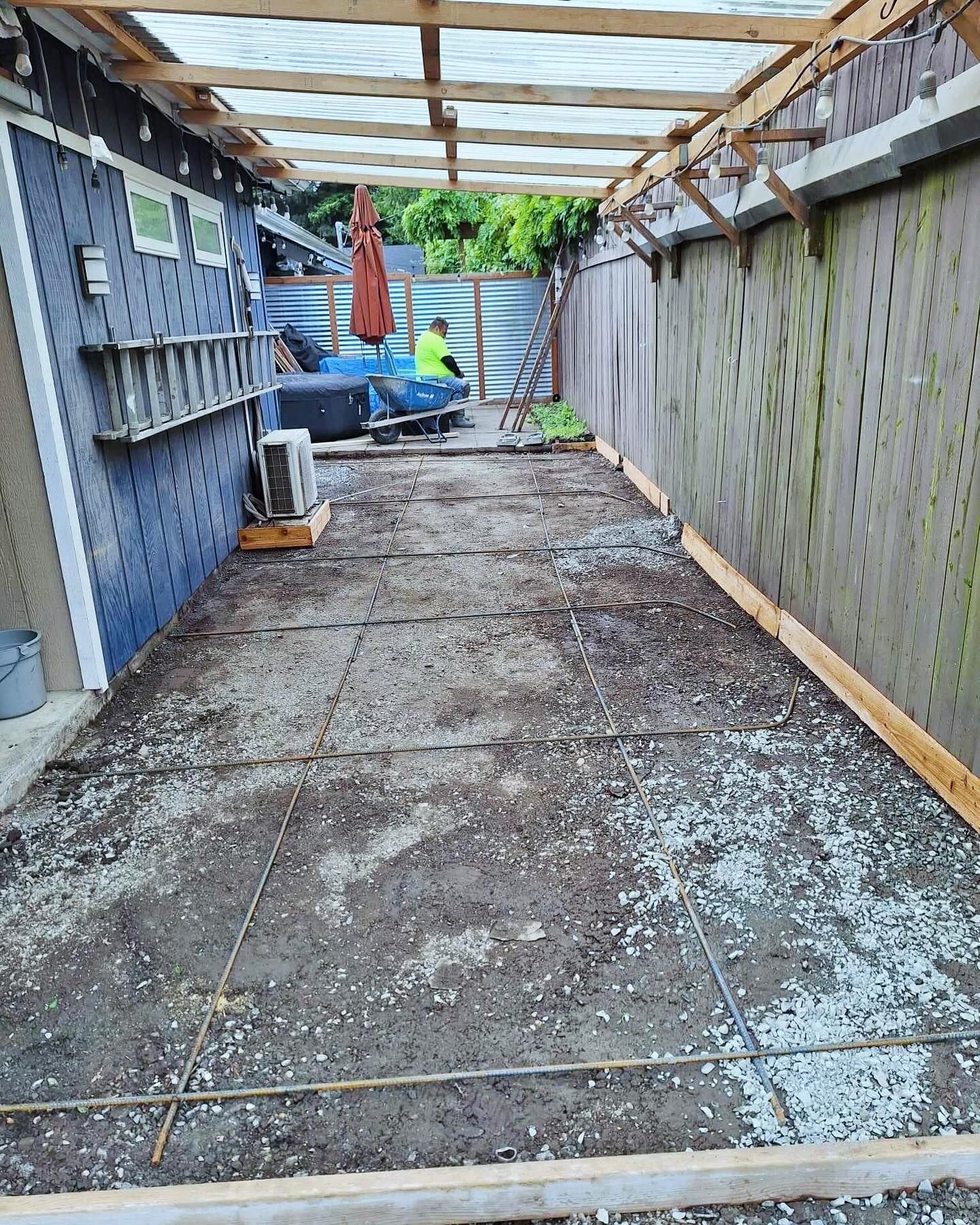 A concrete patio under construction with rebar and wooden frame, between a house and fence.