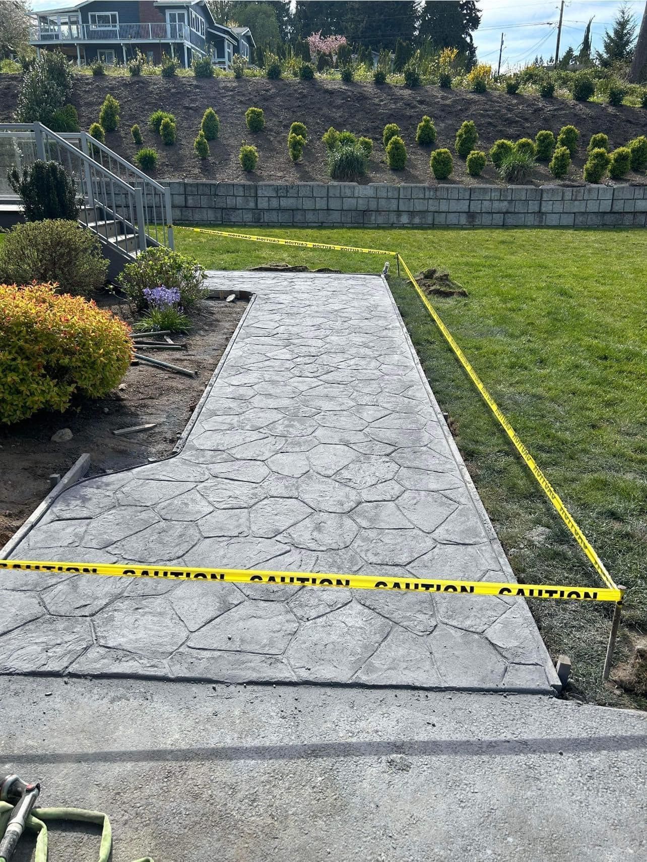 Gray textured concrete pathway under construction, caution tape, green lawn, retaining wall with small trees.
