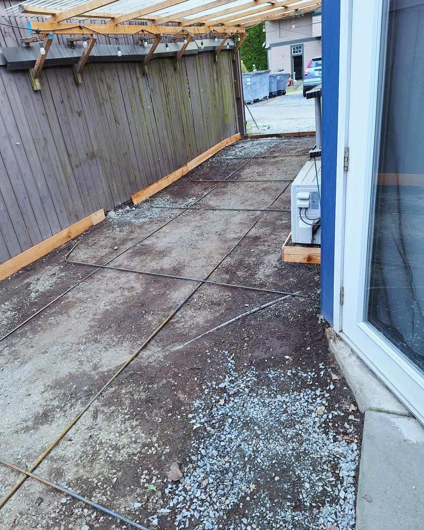 A weathered patio with exposed rebar, next to a wooden fence and a blue-trimmed wall with a window.