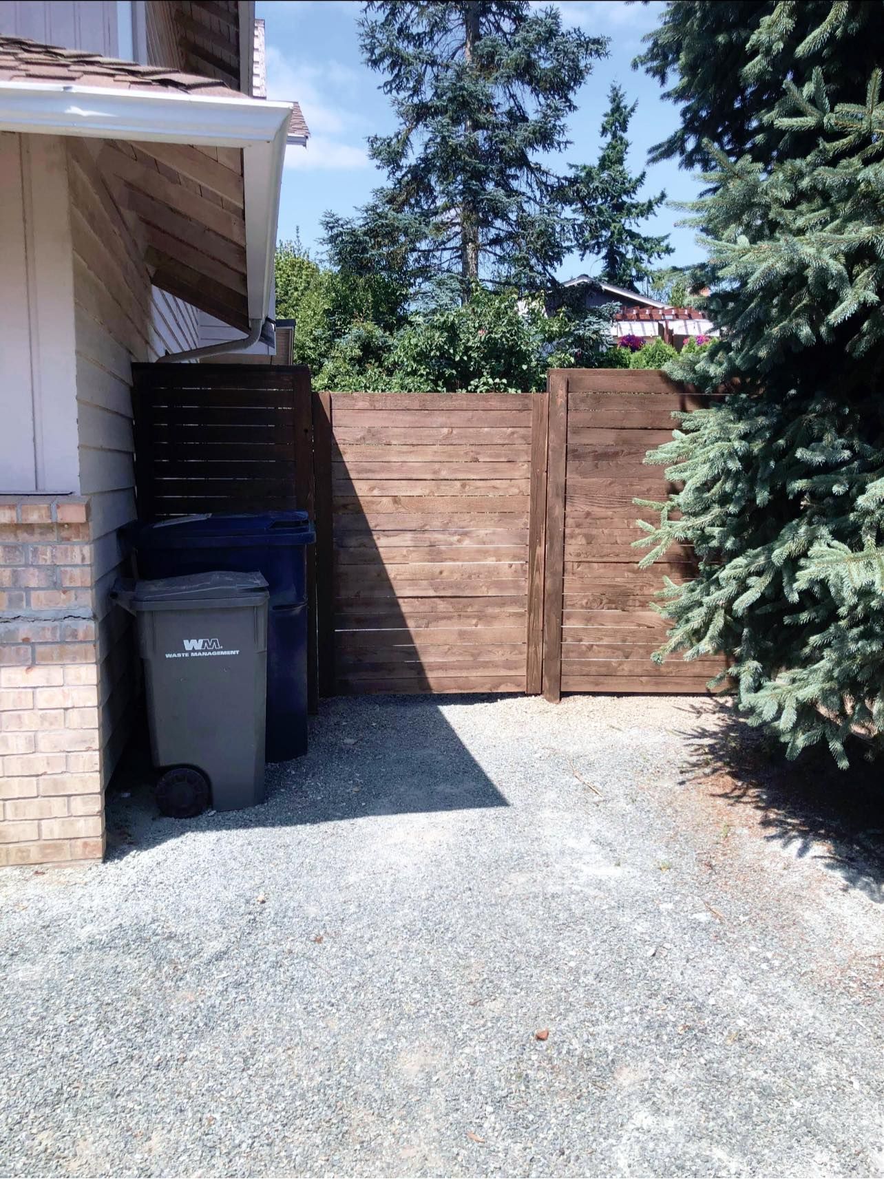 Gravel driveway with trash cans next to a brown wooden fence, next to a white house with a green tree.