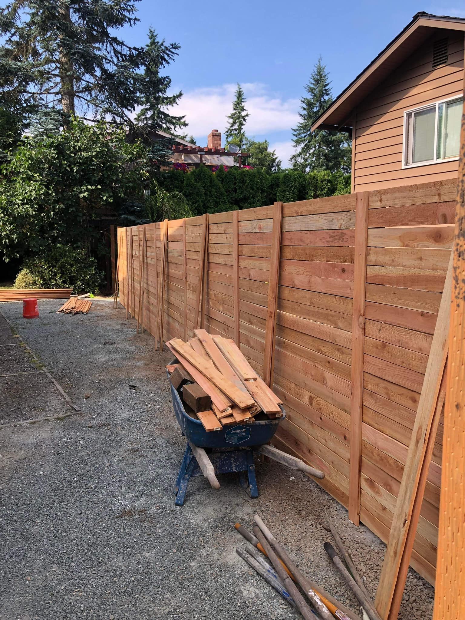 Wooden fence being constructed, with wood and a wheelbarrow in a gravel area.