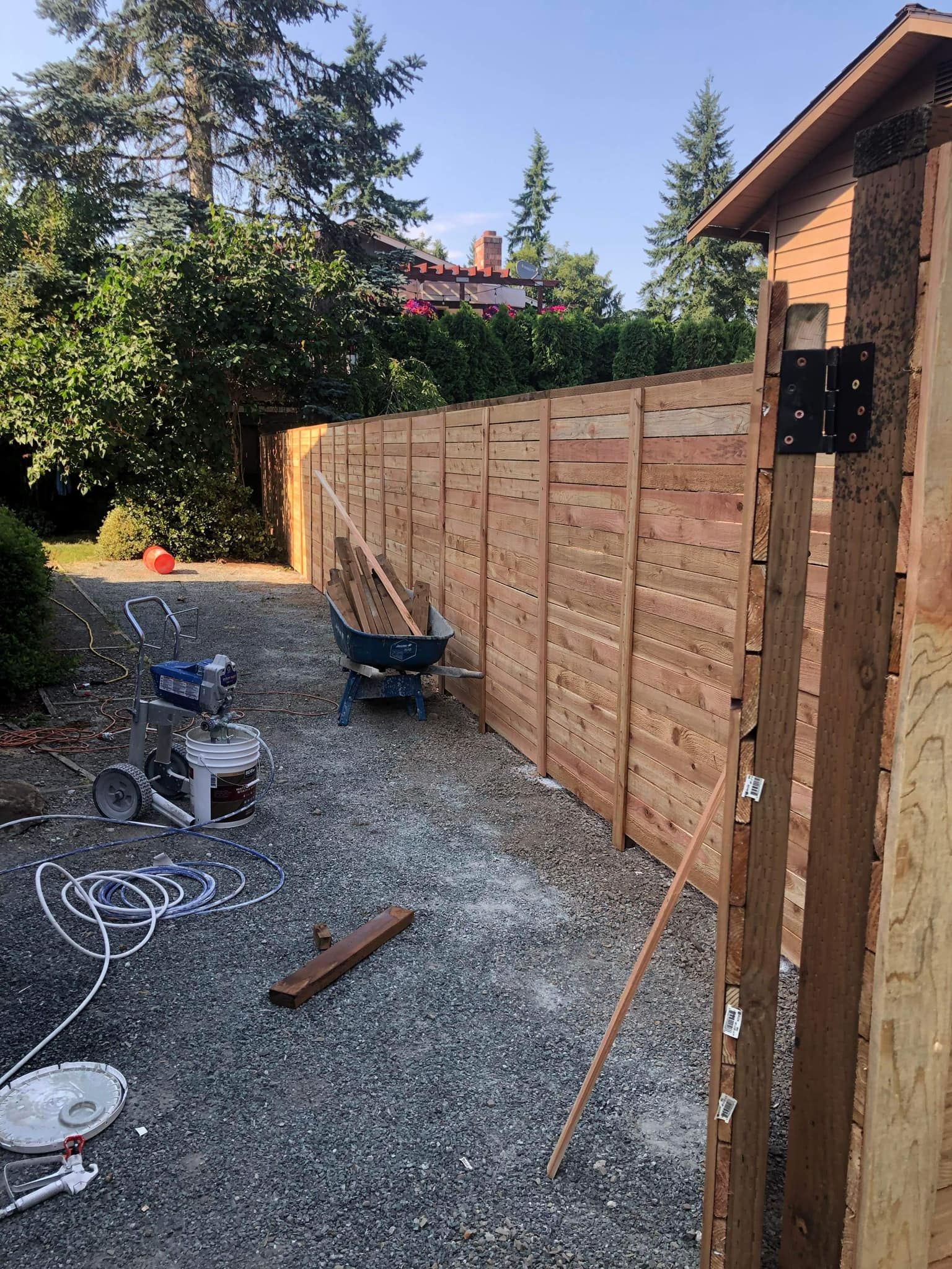 A wooden fence under construction in a gravel driveway. Tools and wheelbarrow nearby.