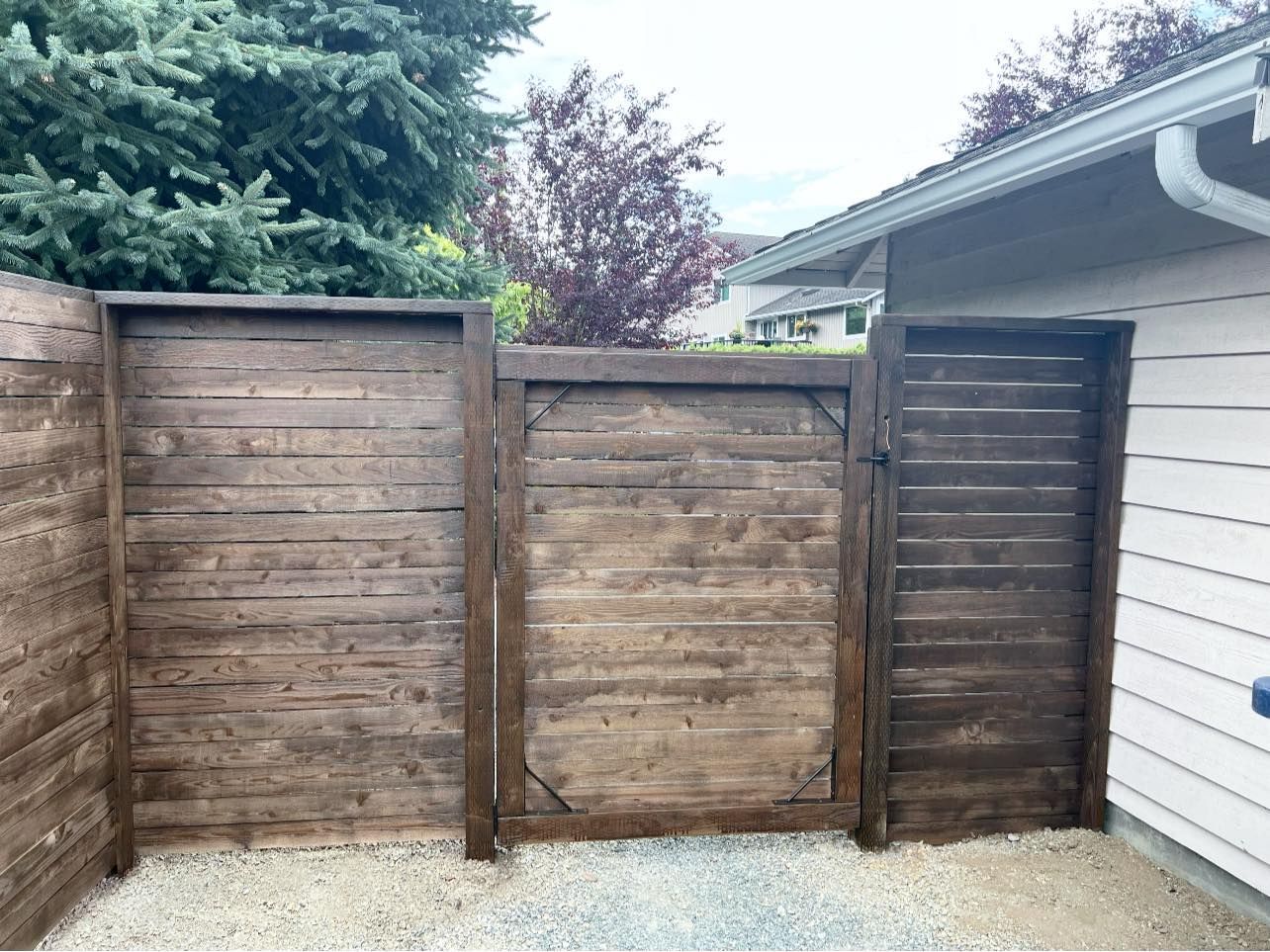Wooden privacy fence with gate beside a white house and gravel ground.