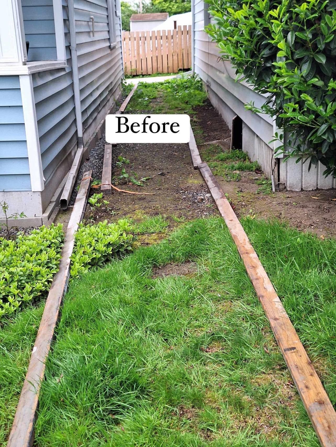 Narrow, unkempt yard between two houses. Wood borders define the path, with grass and weeds present. 