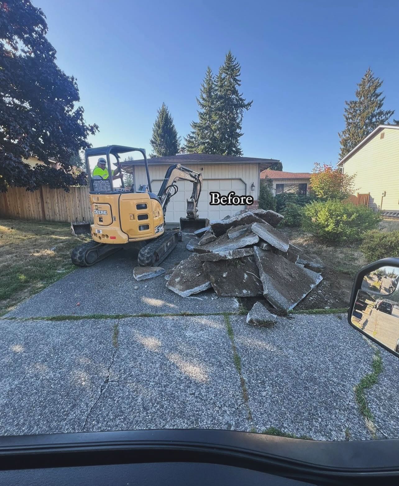 Yellow excavator removing concrete slabs from a driveway, next to a garage.