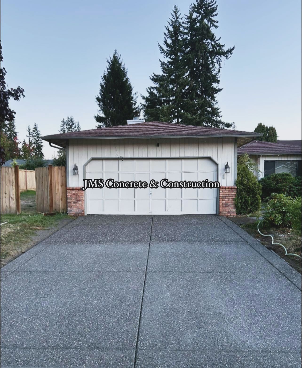 Concrete driveway leading to a white garage with brick accents and a brown roof. Two trees are in the background.