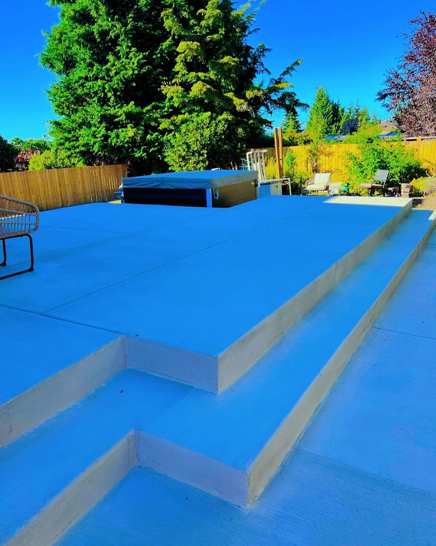 Concrete patio with steps, hot tub, and bamboo fence, against a backdrop of trees under a blue sky.