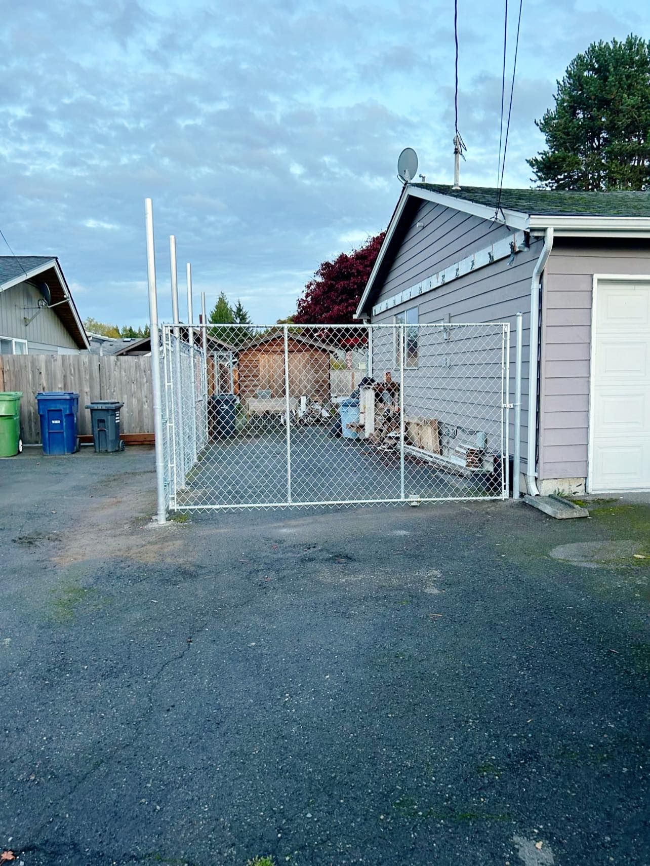 Chain-link fence encloses a driveway area next to a gray house, with garbage cans visible on the left.