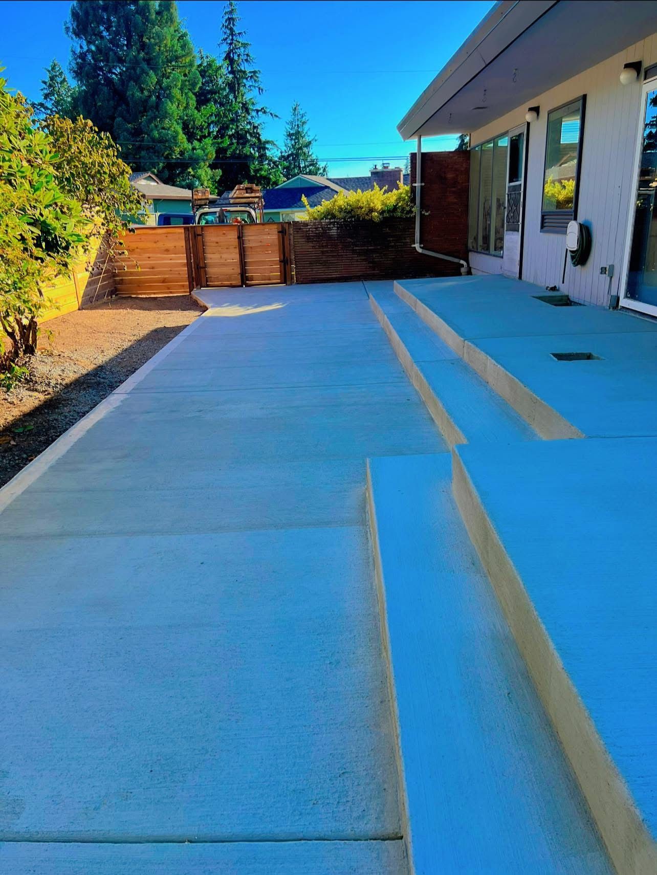 Exterior view of a newly poured concrete patio with steps leading to a wooden fence and gate, a blue sky is visible.