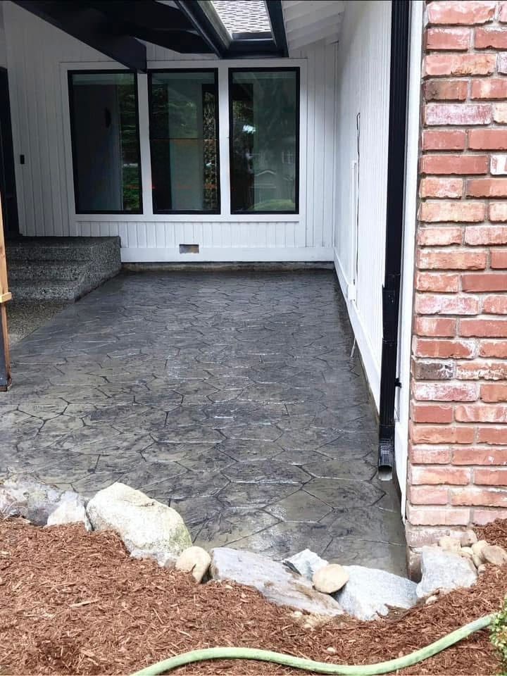 Patio with stamped concrete, white siding, and brick wall. Rocks and dirt border the patio's edge.