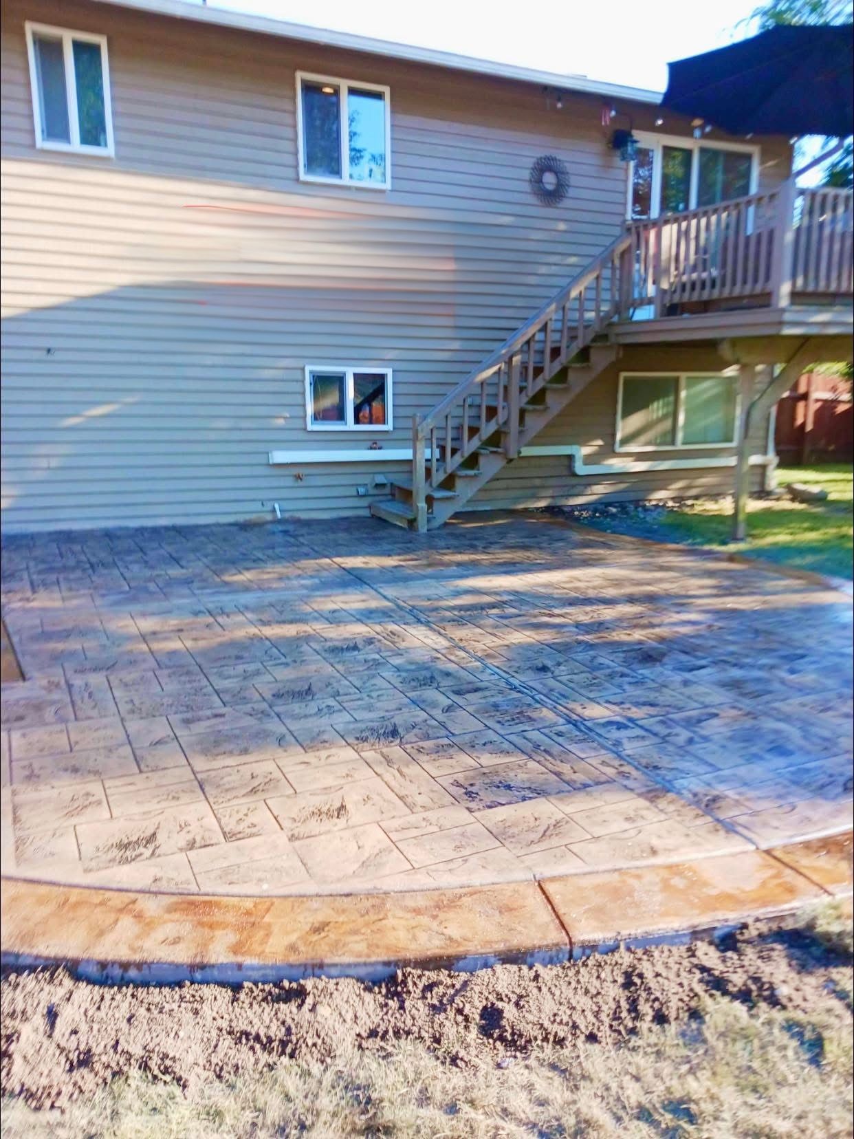 Stamped concrete patio with brown border next to a two-story house with a wooden deck and stairs.