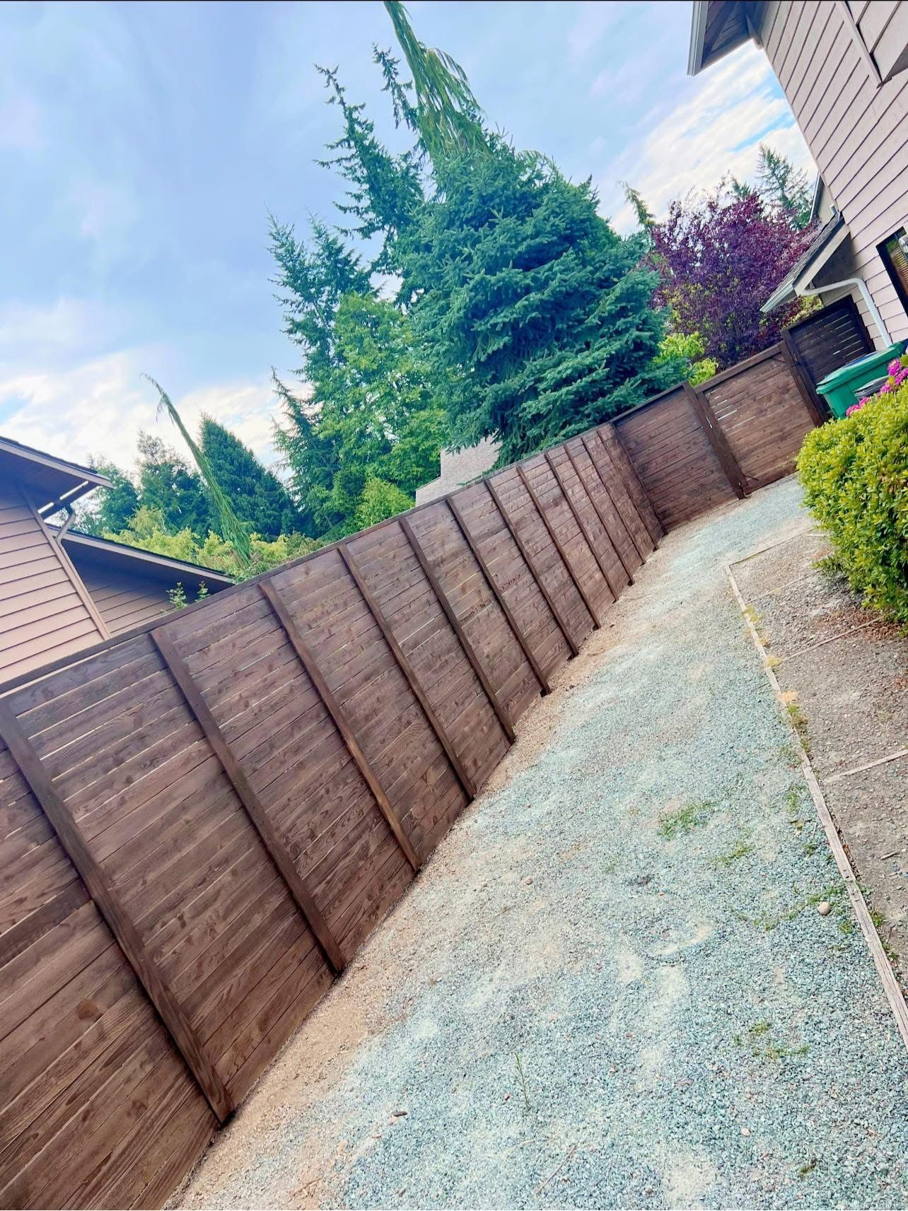 Brown wooden fence along a gravel path, with trees and a building in the background.
