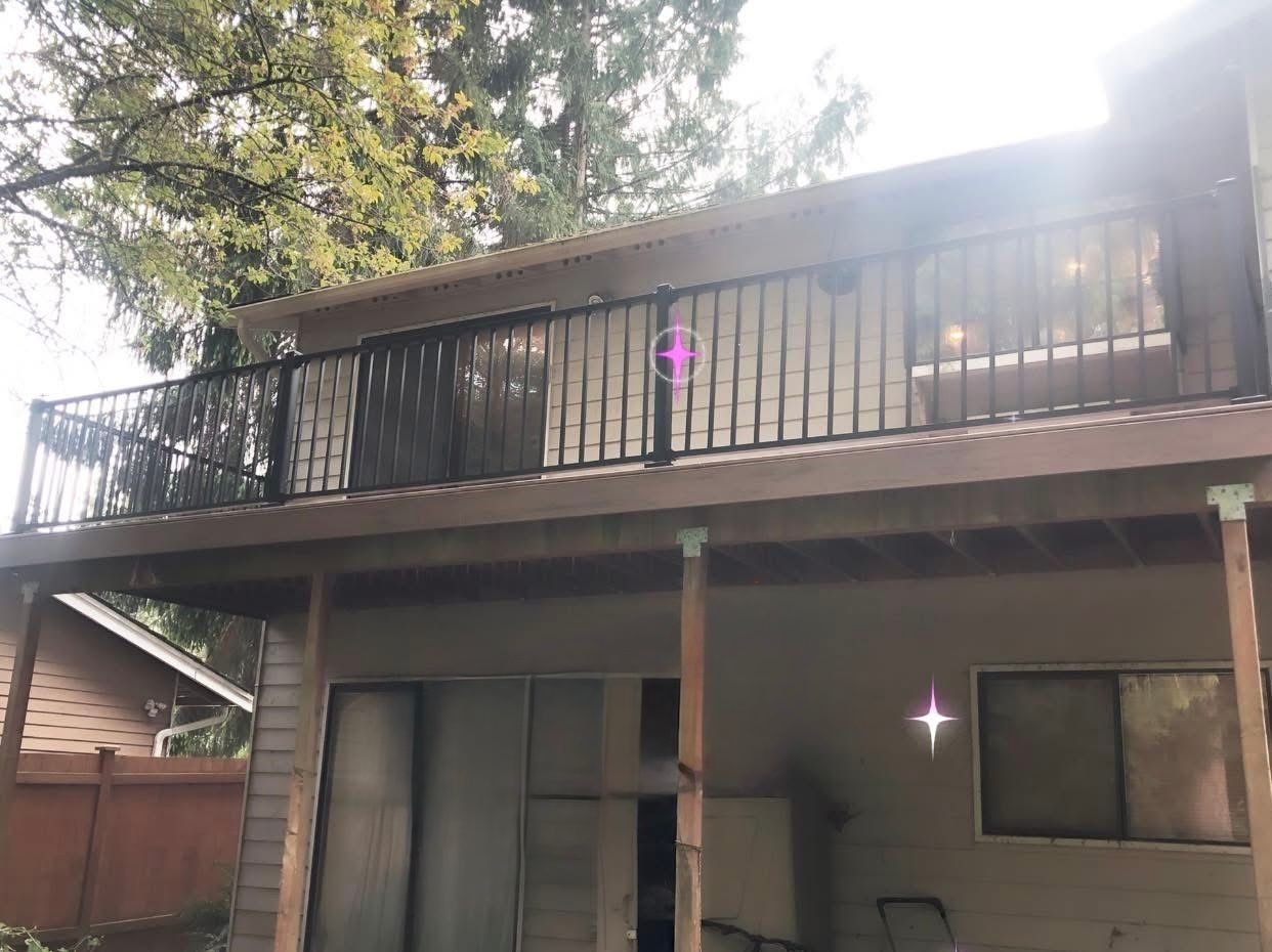 Two-story house with a wooden deck and black metal railing, under a tree canopy.