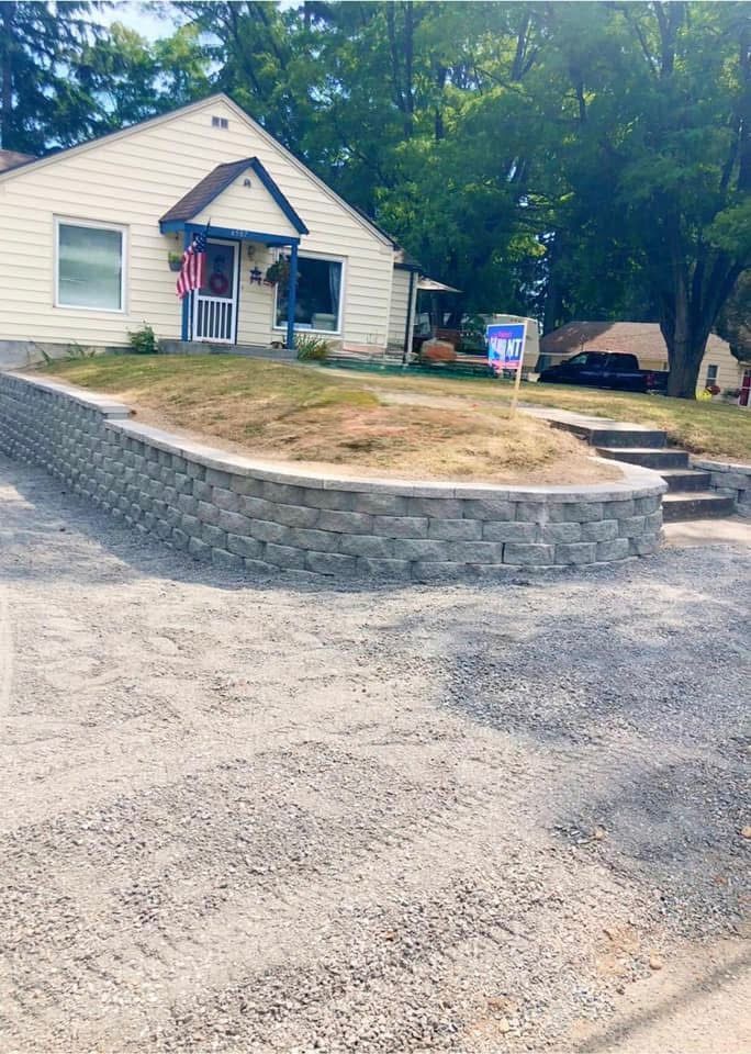 Gray retaining wall in front of a light yellow house with a porch and steps leading up to the front door.