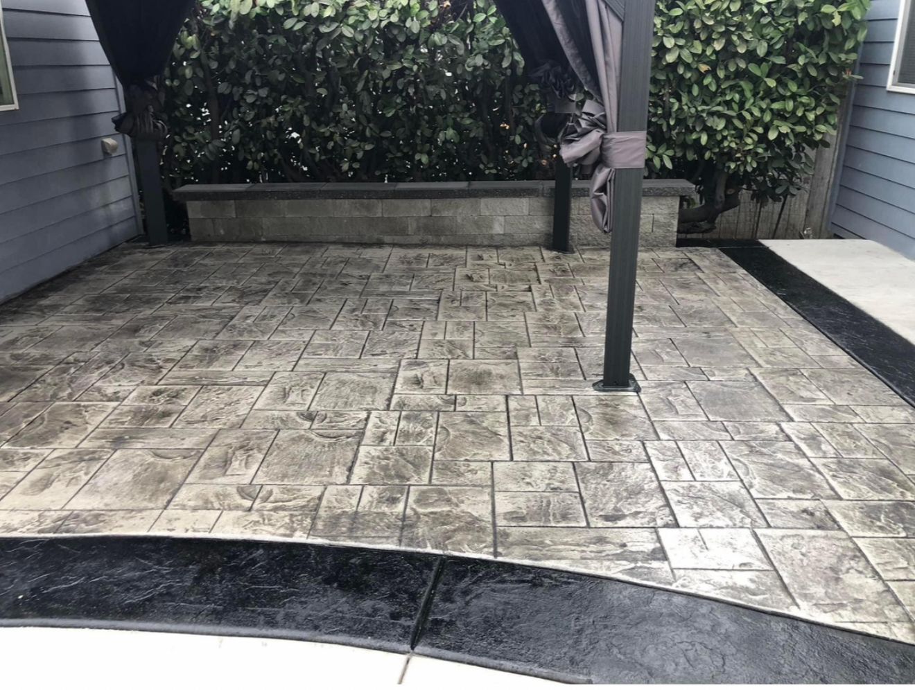 Stamped concrete patio with a dark border, a gazebo post, and a low wall with foliage in the background.
