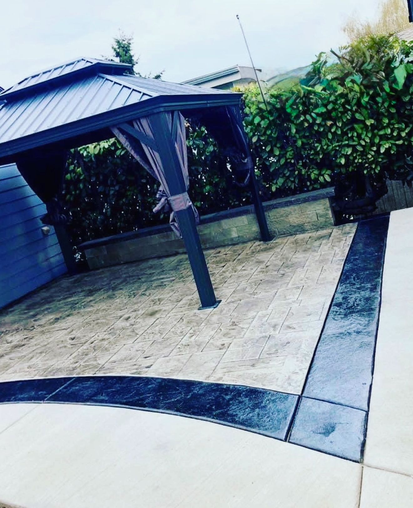 Patio with a dark-roofed gazebo, stamped concrete, and a surrounding border. Green hedge in the background.