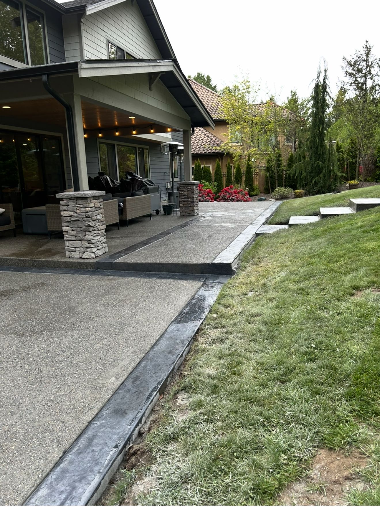 Concrete patio with stone columns, edged with a dark border, next to a grassy hill and a house.