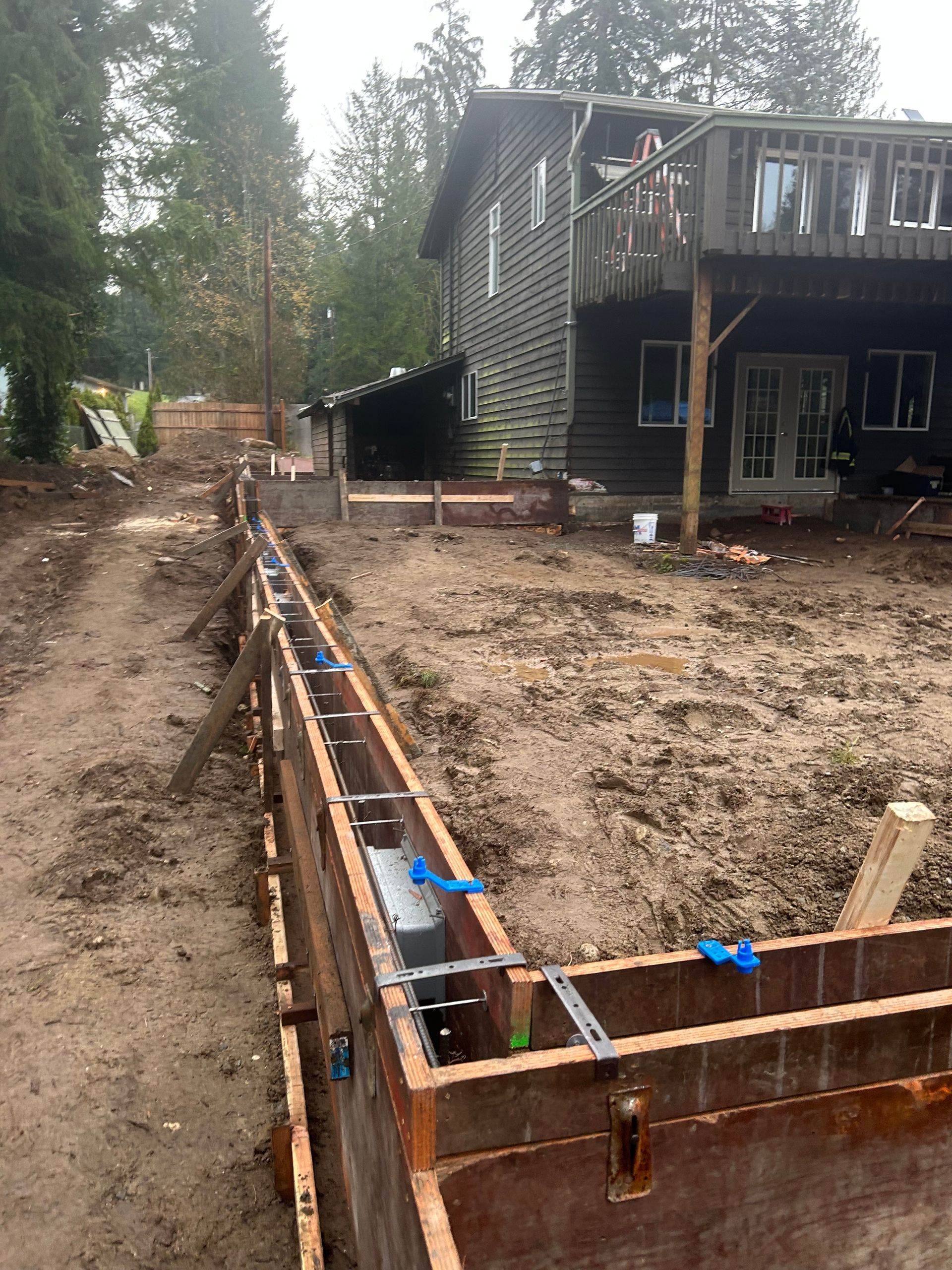Wooden forms for a retaining wall under construction in a muddy yard, near a house with a deck.