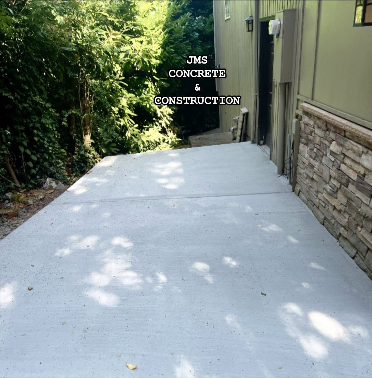A newly poured concrete patio alongside a building with stone veneer and lush green foliage.
