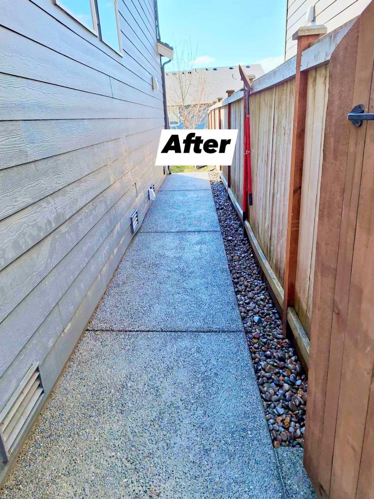 Narrow concrete walkway between a light gray house and a wooden fence with gravel bed.